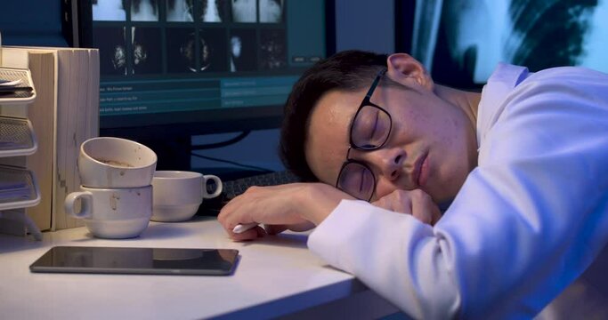 Exhausted overload asian male doctor is falling asleep on desk with monitors showing x-ray film. Sleepy tired alone physician man dozing off while holding stylus in one hand at late night. Dolly shot.