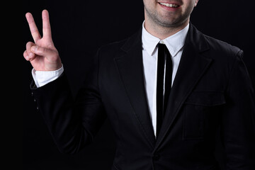 Unrecognizable young man in a black suit, smiling, doing victory sign with two fingers. Isolated on black background. No face. Studio.