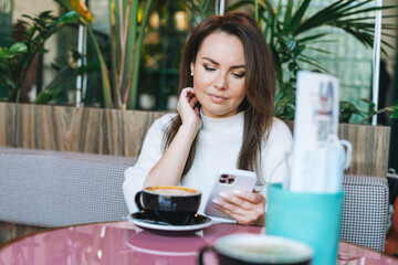Attractive young brunette smiling woman in white casual dress with cup of coffee using mobile phone in cafe