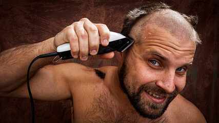 Caucasian man trying to shave his head with an electric razor. A brutal bald man holds a razor in...