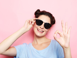 Portrait of a happy young brunette woman wearing sunglasses showing ok gesture over pink background
