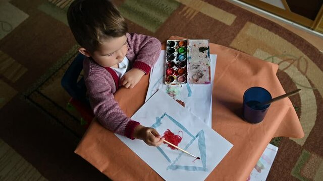 Little son painting cute home using colourful paints and sheet of white paper. Housing construction concept. Top view of a children's desktop, paints, paper, brushes and glass of water
