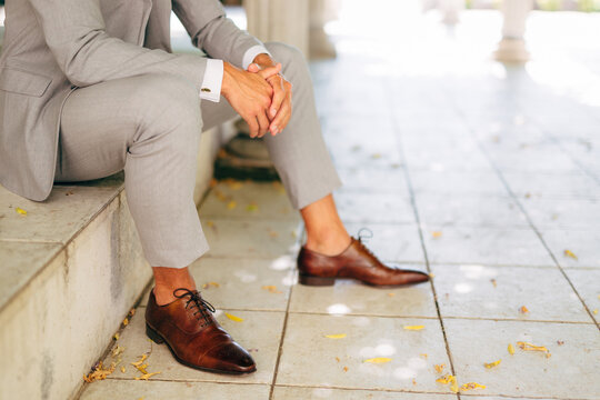 A man in a gray suit and leather brown shoes sits on the step with his hands folded 