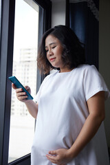 Fototapeta na wymiar Young Asian Pregnant Woman standing near the window and using smart phone for checking information