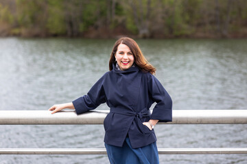 A woman with bright red lipstick on her lips stands at the fence on the river embankment. The woman smiles, the wind blows her long hair.