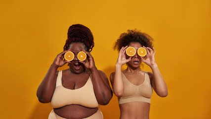 Two african american women in underwear covering their eyes with halves of ripe juicy orange, posing together isolated over orange background