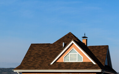 Multi-gable roof of house. Roof window. Flexible roofing material in brown color. Brick, chimney pipes. Background - blue sky.