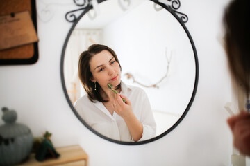 Cute caucasian woman with Jade Massage Roller near mirror ,female brunette does facial massage with roller, gouache. Concept beauty and self-care, skin and face care