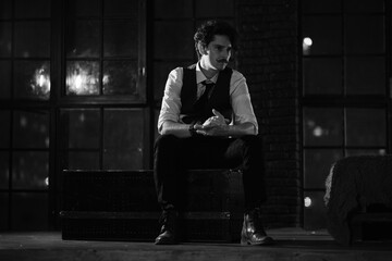 stylish dressed man stands at the window and looks into the distance. pensive man stands at night in a dark room. black and white photography