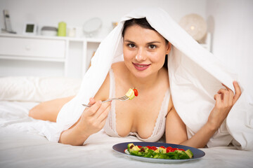 Fototapeta na wymiar Young sexy woman undeer blanket eating vegetable salad from plato in bed at home
