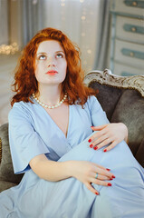 Cheerful red- haired curly young woman with blue eyes and freckles sits in large elegant armchair in studio. Beautiful people. 