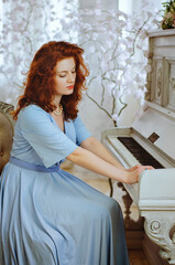 Beautiful red- haired young female pianist with blue eyes and freckles sits near a piano and learning music in studio. The creative process of making music.