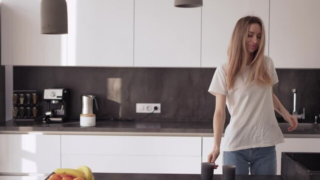 A young woman is dancing in the kitchen and drinking coffee