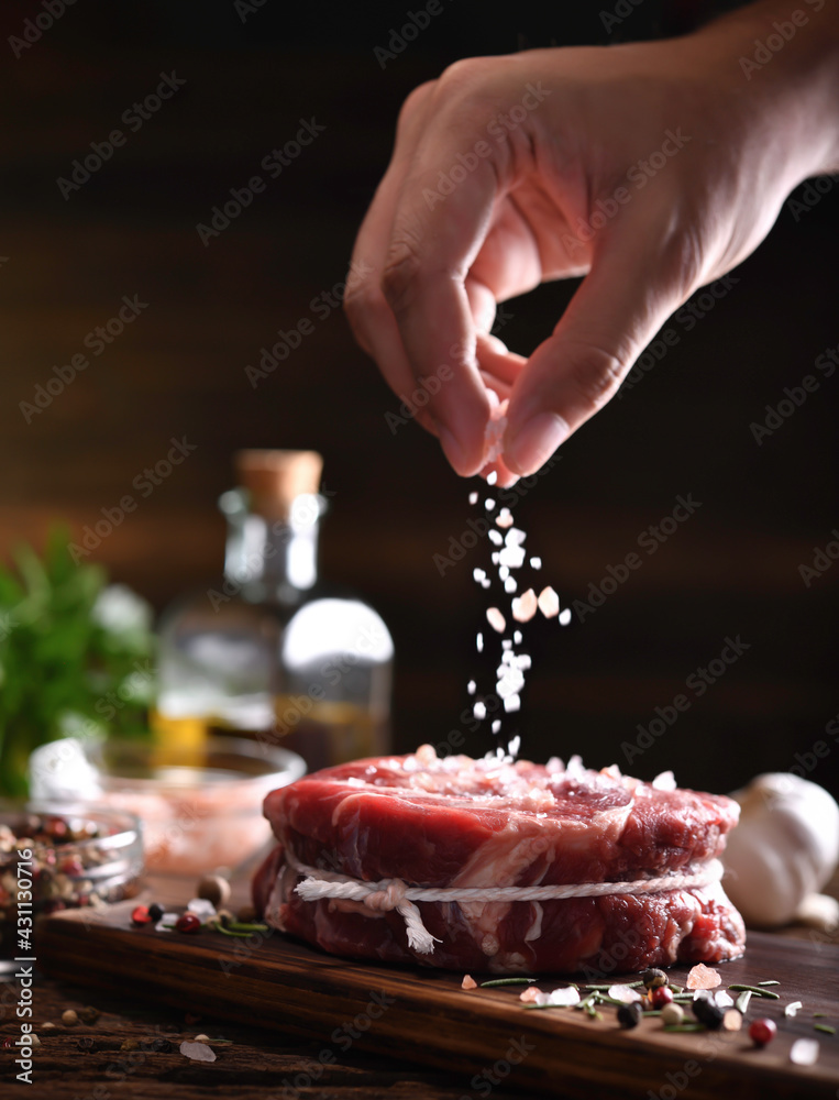 Wall mural Hand sprinkling salt on fresh raw beef meat on a cutting board