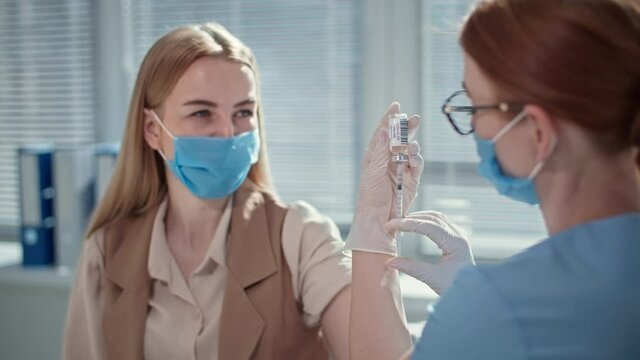 doctor uses hypodermic needle and syringe to put shot of drug as treatment to young female patient wearing medical mask