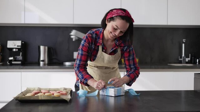 Woman ties a bow on a gift box with makaoons at kitchen