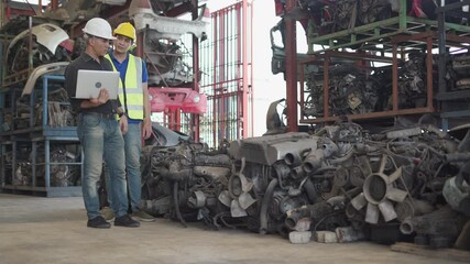 senior engineer using laptop computer checking list old used car engine with young asian worker in machinery parts shop warehouse. old manager find vehicle automobile machine in Recycle motor factory