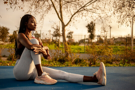 Black woman sitting down on the floor holding her knee and streching her leg. Outdoor activities.