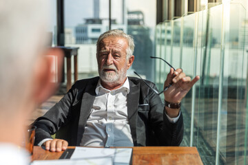 Old business man in his 60th wearing white shirt and black jacket. He is also holding glass in his hand. He sit by the window in a hotel lobby.