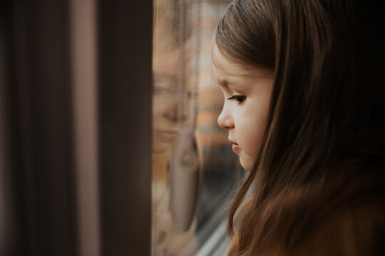 Beautiful little girl smiles and looks out the window. The child looks out the window. A young girl looks out of the window. A portrait of a cheerful kid on the windowsill.