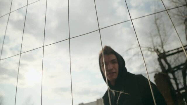 young Asian man in a black hoodie walks up to the fence, pushes it with his hand. Looking into the camera. Aggressive guy. Unrest in the city and rectification of youth.