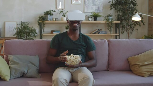 Young cheerful Afro-American man sitting on sofa at home, eating popcorn and smiling while watching movie on VR headset