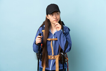 Young Chinese girl with backpack and trekking poles over isolated blue background having doubts and thinking
