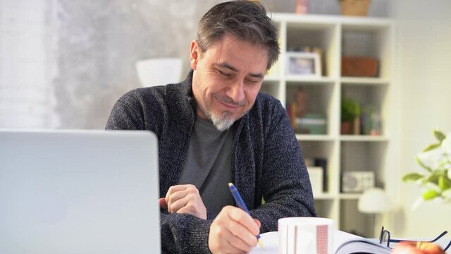Middle aged man sitting at desk in morning at home, reading news on laptop. Businessman working in home office with computer, smiling.
