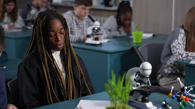 Close-up of upset dark-skinned girl student with afro-braids doing nothing during chemistry lesson being stressed. Dyslexic teenage pupil sitting blankly at school desk in classroom