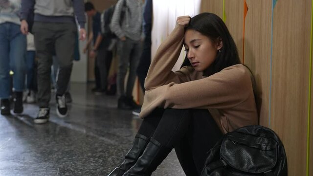 Close-up of upset chinese schoolgirl sitting on floor in school corridor while other students passing by ignoring her. Indifferent classmates leaving despaired girl out and do not pay attention to her