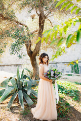 Lovely bride in a beige dress stands against a tree background with a gorgeous bouquet of flowers in her hands