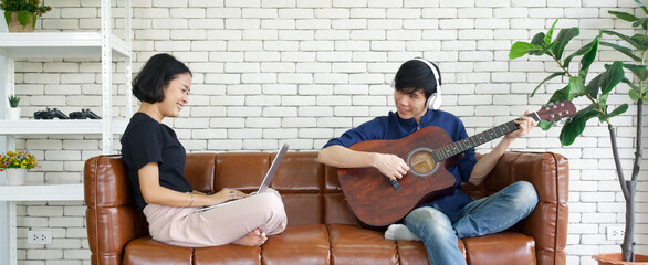 Young lovers spend time together on holiday in the living room. Asian man with headphones playing an acoustic guitar while the girl use laptop computer to finish pending work.