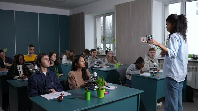 Mixed race female teacher using model while asking multi-ethnic high school students about molecular structure of substance. Multiracial classmates raising hands to answer teacher's question in class