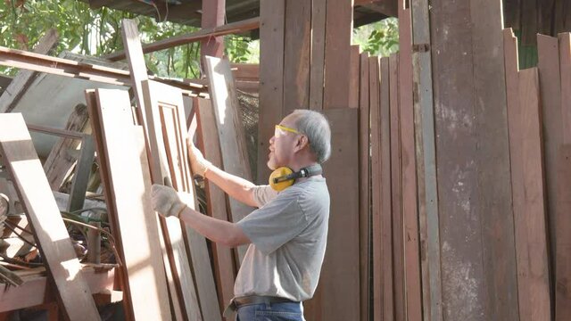 Old man carpenter carefully looking and inspecting wood plank in the factory.
