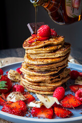 Pouring maple syrup on healthy banana oatmeal pancakes stack with strawberries, bananas, raspberries and chocolate. Easy gluten free morning breakfast or brunch. Delicious pancakes heap, maple drizzle