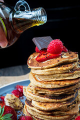 Pouring maple syrup on healthy banana oatmeal pancakes stack with strawberries, bananas, raspberries and chocolate. Easy gluten free morning breakfast or brunch. Delicious pancakes heap, maple drizzle
