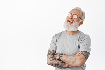 Image of candid senior man in glasses, laughing with arms crossed on chest, looking left aside with joyful relaxed face expression, standing over white background