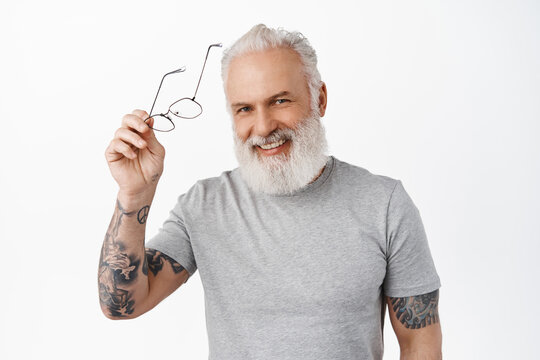 Handsome Senior Man With Tattoos, Take Off Glasses, Laughing And Smiling Happy. Stylish Hipster Old Guy Looking Cheerful At Camera, Standing Over White Background