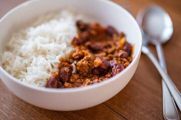 Close up and selective focus of meat chilli concarne and white rice in a white bowl on a wooden surface with intentional shallow depth of field and bokeh