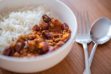 Close up and selective focus of meat chilli concarne and white rice in a white bowl on a wooden surface with intentional shallow depth of field and bokeh