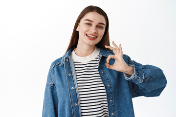 Okay you alright. Smiling happy girl shows OK sign in approval, give positive answer, say yes, nod and recommend event, guarantee quality, standing over white background