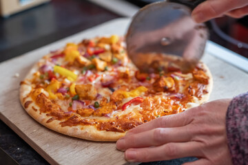 Close up and selective focus of a home cooked thin crust sweet chilli chicken pizza on a wooden cutting board being sliced with a pizza roller by an unidentifiable woman