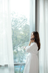 Fototapeta premium Young Asian woman in bathrobe standing at apartment window, drinking morning coffee and talking on phone