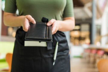 Cropped image of woman waitress in apron hold out bill folder to cafe visitor of cafe or restaurant.