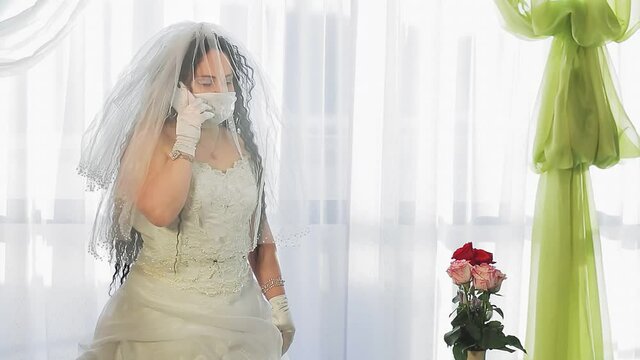 A Jewish bride in a wedding dress and a veil in a medical mask in the synagogue hall before the hupap ceremony accepts congratulations and speaks on the phone.