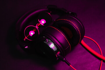 Fototapeta na wymiar large DJ headphones with a red cable lie on the table along with round-shaped sun glasses and are illuminated by a purple light