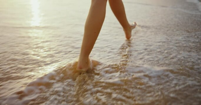 Woman walking down the beach at sunset, close up of feat in the sand and waves washing up the beach