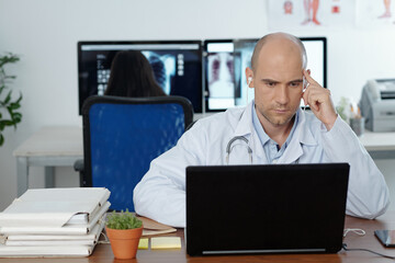 Serious concentrated general practitioner wearing earphones when watching webinar on laptop, his colleague is working in background