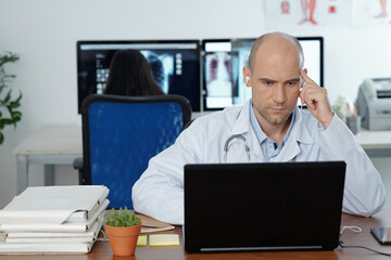 Fototapeta premium Serious concentrated general practitioner wearing earphones when watching webinar on laptop, his colleague is working in background