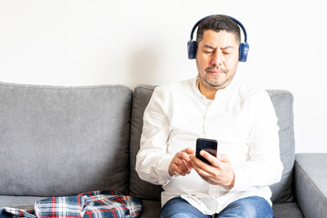 Hispanic man playing music from his mobile phone listening with his wireless headphones on his comfortable sofa at home.